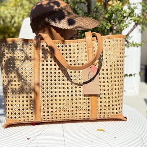 May include: A rectangular tote bag crafted from light tan woven cane webbing featuring a repeating octagonal pattern. The bag includes tan leather trim along the top, base, and vertical straps, with a rectangular cardboard tag attached to the front.