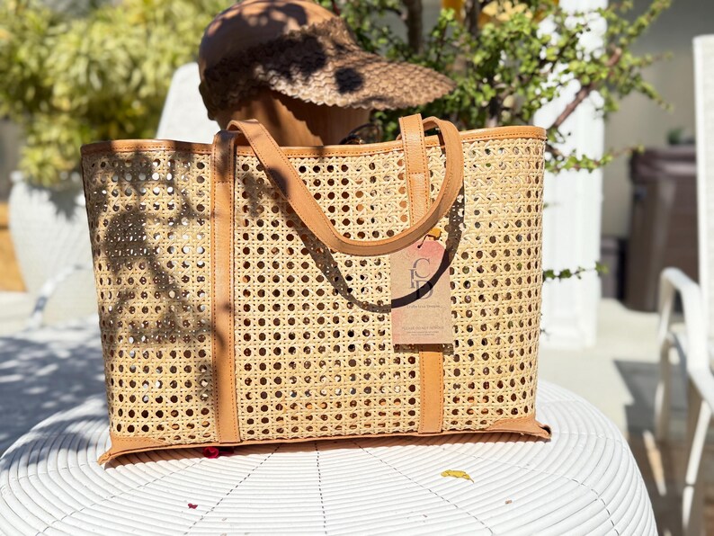 May include: A rectangular tote bag crafted from light tan woven cane webbing featuring a repeating octagonal pattern. The bag includes tan leather trim along the top, base, and vertical straps, with a rectangular cardboard tag attached to the front.