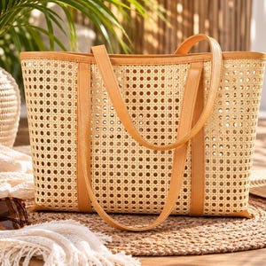 May include: A rectangular tote bag crafted from light beige woven rattan cane webbing, featuring tan faux leather trim along the top edge and vertical side panels, complemented by two matching tan faux leather shoulder straps.