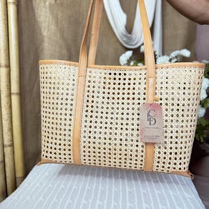 May include: A rectangular tote bag crafted from light beige woven cane webbing with tan leather trim and two matching leather shoulder straps. A rectangular tag with the letters CD and text is attached to the front of the bag by a thin cord.