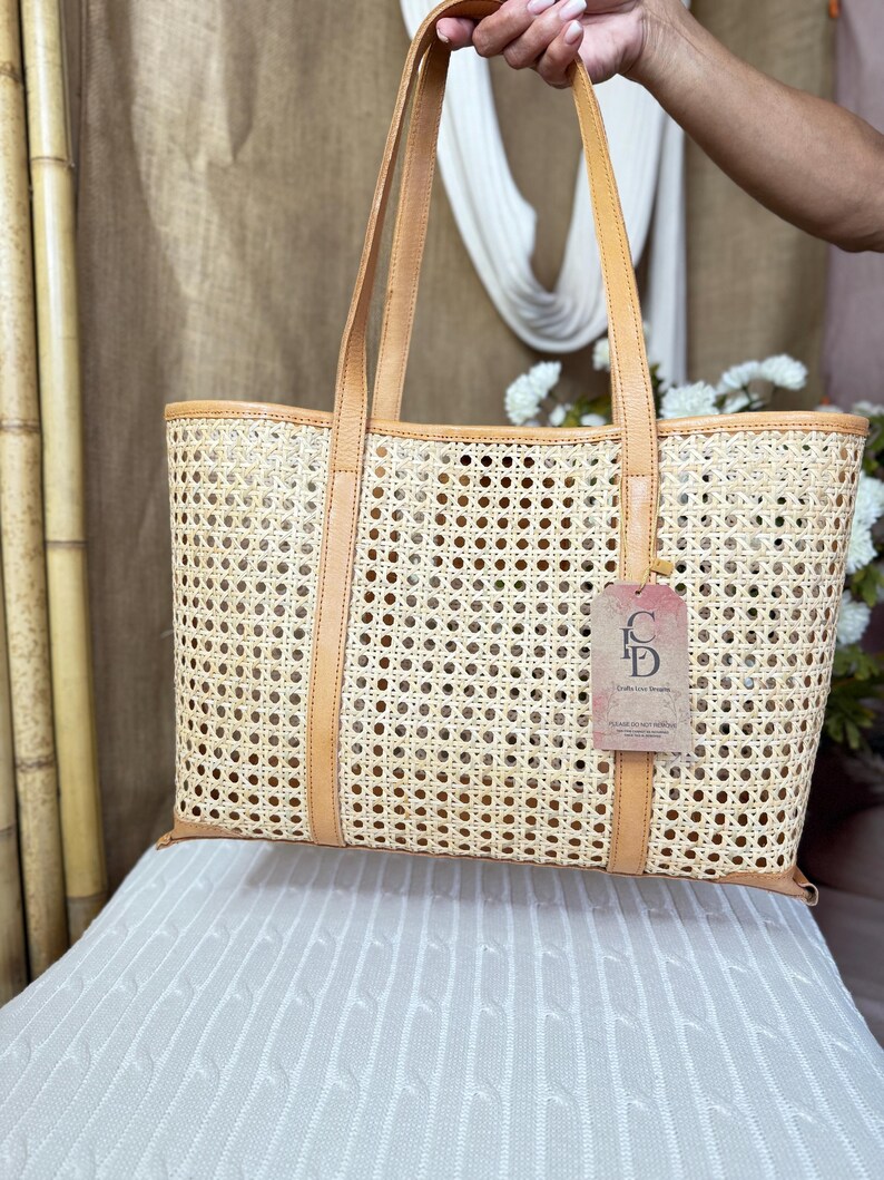 May include: A rectangular tote bag crafted from light beige woven cane webbing with tan leather trim and two matching leather shoulder straps. A rectangular tag with the letters CD and text is attached to the front of the bag by a thin cord.