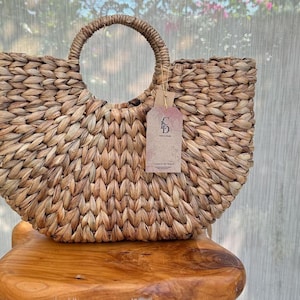 A woven handbag with a round handle and a tag attached, placed on a wooden stool against a backdrop of a white curtain.