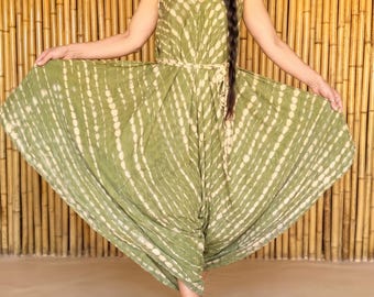 May include: An olive green jumpsuit with a tie-waist and a V-neck, patterned with white dotted stripes. The model wears a wide-brimmed straw hat and sunglasses. The background is a bamboo wall. The jumpsuit is a flowing design.