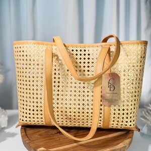 A tan woven tote bag with a handle and a tag attached, placed on a wooden stand against a blue curtain background.
