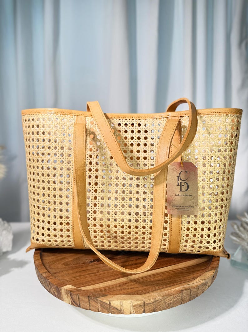 A tan woven tote bag with a handle and a tag attached, placed on a wooden stand against a blue curtain background.