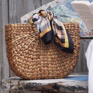 A woven straw bag with a plaid scarf tied around it, placed on a wooden surface.