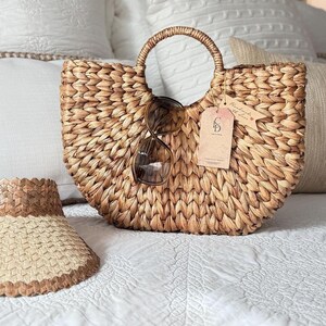 A woven straw handbag with a tag attached, a beige sun visor, and a pair of sunglasses are arranged on a white bedspread.