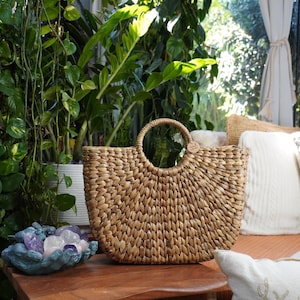 A woven straw handbag sits on a wooden table, surrounded by lush greenery and a cozy seating area with pillows and a bird-themed pillow.