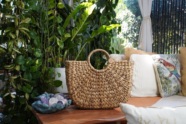 A woven straw handbag sits on a wooden table, surrounded by lush greenery and a cozy seating area with pillows and a bird-themed pillow.