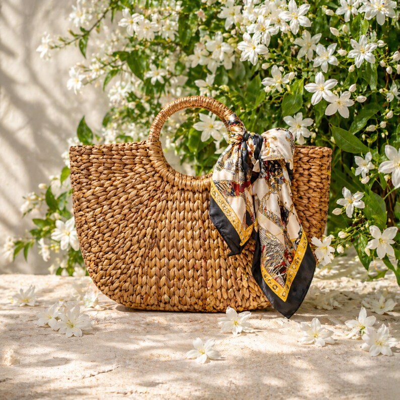 A woven straw handbag with a black and gold patterned scarf draped over it, placed on a white surface with white flowers in the background.