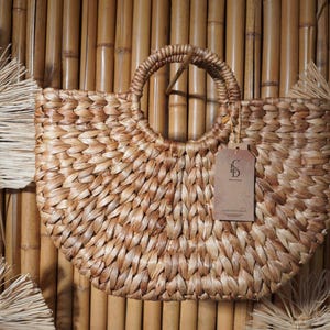 A woven straw bag with a handle, hanging on a bamboo wall.