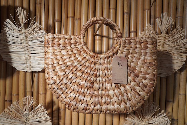 A woven straw bag with a handle, hanging on a bamboo wall.