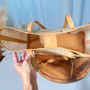 A hand holding a woven straw bag with a wooden base, against a blue background.