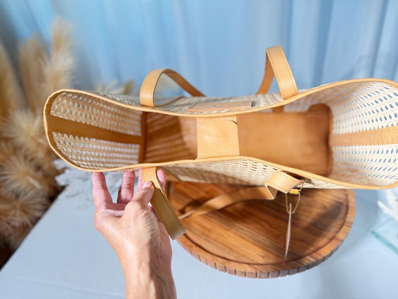 A hand holding a woven straw bag with a wooden base, against a blue background.