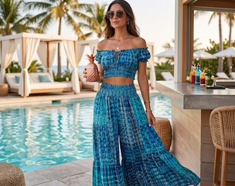 May include: A blue tie-dye two-piece outfit, comprising an off-the-shoulder crop top and wide-leg trousers. The outfit is accessorized with sunglasses and a pineapple-shaped drink. The background includes a pool, palm trees, and a bar.