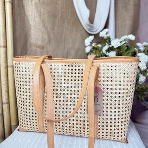 May include: A rectangular tote bag crafted from light beige woven cane webbing, featuring tan leather trim along the top edge and base. The bag includes two long tan leather shoulder straps and a small rectangular paper tag attached to the side.