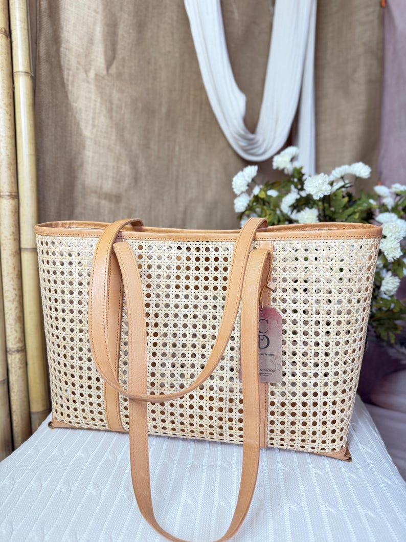 May include: A rectangular tote bag crafted from light beige woven cane webbing, featuring tan leather trim along the top edge and base. The bag includes two long tan leather shoulder straps and a small rectangular paper tag attached to the side.