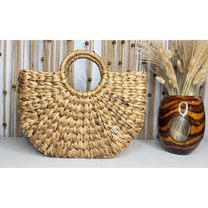 a woven straw bag with a round handle, placed next to a vase filled with dried flowers, creating a rustic and natural aesthetic.