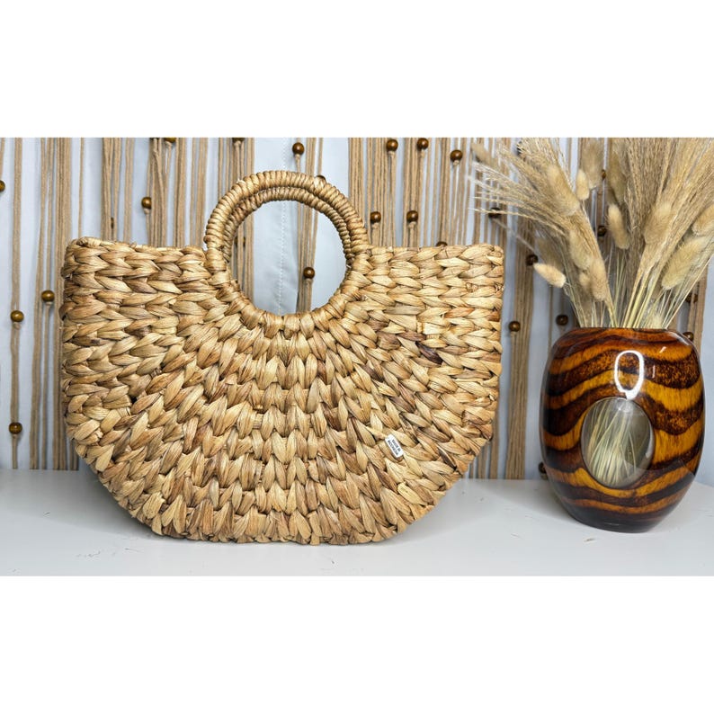 a woven straw bag with a round handle, placed next to a vase filled with dried flowers, creating a rustic and natural aesthetic.
