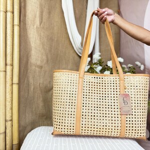 May include: A rectangular tote bag crafted from light tan woven cane webbing with smooth tan leather trim and dual shoulder straps. A rectangular paper tag with the letters CD is attached to the front of the bag by a thin string.
