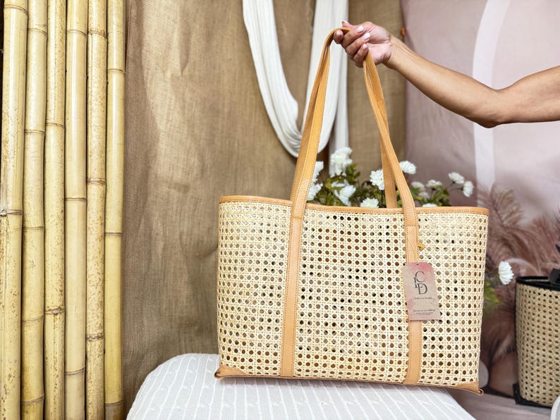 May include: A rectangular tote bag crafted from light tan woven cane webbing with smooth tan leather trim and dual shoulder straps. A rectangular paper tag with the letters CD is attached to the front of the bag by a thin string.