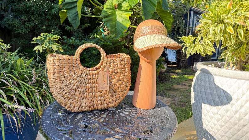 a woven straw handbag and a straw hat placed on a metal table, surrounded by potted plants and greenery.