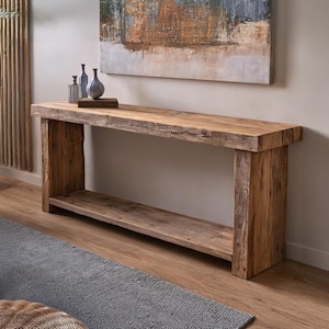 May include: A rustic, rectangular wooden console table with a lower shelf. The table is made of light brown, weathered wood. Decorative vases and a painting hang above the table. A gray rug and woven ottomans are in the foreground.