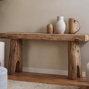 May include: A reclaimed wooden console table with a natural, weathered appearance. The table has a thick, rectangular top supported by two solid, tree-trunk-style legs. Three decorative ceramic items, including a vase and a mug, are displayed on the table's surface.