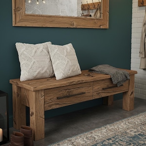 Può includere: Panca in legno rustico con due cassetti, con finitura anticata. Due cuscini color crema a maglia a treccia e una coperta grigia sopra. La panca fa parte di una collezione di arredamento per la casa.