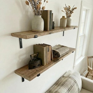 Può includere: Due mensole rustiche in legno fissate a un muro bianco, ciascuna sostenuta da staffe in metallo nero. La mensola superiore contiene un vaso con fiori secchi e libri. La mensola inferiore mostra libri, un ferro da stiro vintage e una coperta piegata.