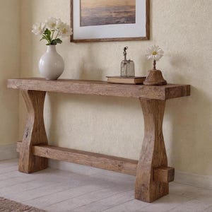 Peut inclure: Une console rustique en bois avec une finition naturelle. La table a un dessus rectangulaire épais et des pieds incurvés. Un vase blanc avec des fleurs et des objets décoratifs sont posés sur la table.