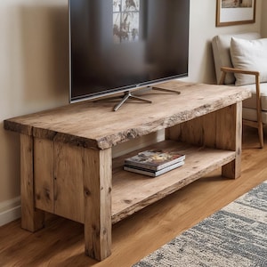 May include: A rustic, wooden TV stand with a natural, unfinished look. The stand features a lower shelf and thick, sturdy legs. The wood has visible knots and grain, adding to its unique character. A large TV is placed on top of the stand.