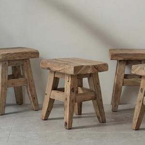 Puede incluir: Cuatro taburetes rústicos de madera con asientos cuadrados y patas anguladas. Los taburetes son de madera marrón claro y tienen un aspecto natural y envejecido. Están colocados sobre un suelo de baldosas grises.