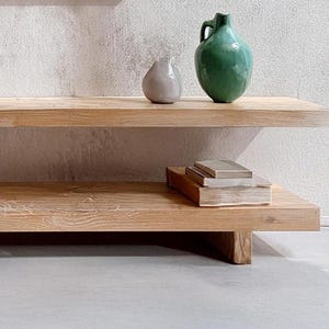 May include: A two-tiered wooden shelf with a light wood finish. The top shelf has a green ceramic pitcher and a gray ceramic pear. The bottom shelf has a stack of books.