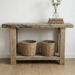 May include: A rustic wooden console table with two woven baskets on the lower shelf. The table is made of weathered wood and has a natural, unfinished look. A glass vase with flowers and a stack of books sit on top.