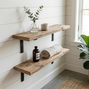 May include: Two rustic wooden shelves with black metal brackets are mounted on a white wall. The top shelf holds a clear glass vase with greenery and a small beige ceramic cup. The bottom shelf displays a rolled towel and a brown bottle.