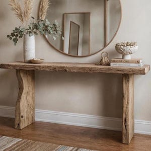 Peut inclure: Une console en bois rustique avec un dessus à bord brut et deux pieds épais de forme organique. La table est ornée d'un vase de plantes séchées, d'un bol décoratif, de livres et d'un miroir rond. Les couleurs sont neutres avec des tons de bois naturels.