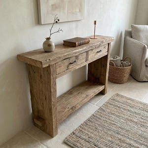 Può includere: Una consolle rustica in legno con due cassetti e un ripiano inferiore. Il tavolo è realizzato in legno di recupero e ha una finitura naturale. Un piccolo vaso con un rametto, un libro e un portacandele sono sopra. Un tappeto intrecciato è sul pavimento. Il tavolo è lungo circa 120 cm.