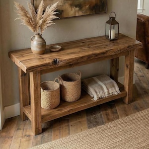 May include: A rustic wooden console table with a natural live edge top and a lower shelf. The table holds a ceramic vase with dried pampas grass, a small bowl, and a metal lantern. Two woven baskets and a folded beige throw blanket sit below.