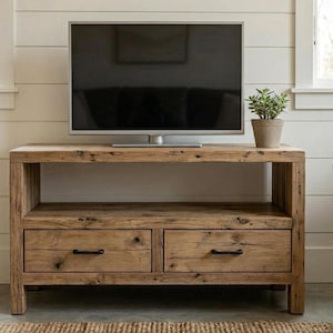 Può includere: Mobile TV rustico in legno chiaro con due cassetti e un ripiano inferiore. Il mobile è realizzato in legno invecchiato e ha maniglie nere. Un televisore a schermo piatto è posizionato sopra e una piccola pianta in vaso è a destra.