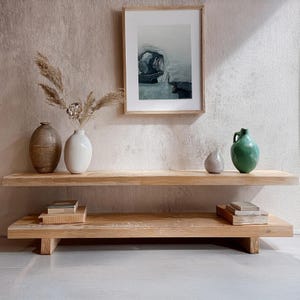 May include: A two-tiered wooden shelf unit with a natural wood finish. The shelf is decorated with three ceramic vases, two of which are white and one is brown. The shelf is also decorated with a framed print of an abstract landscape.