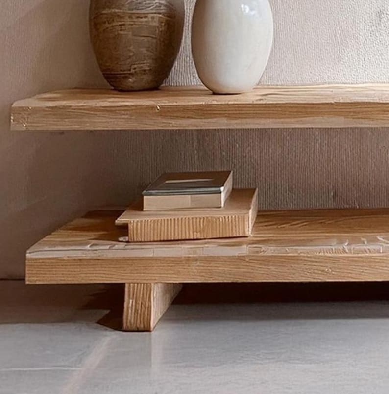 May include: A two-tiered wooden shelf unit with a natural wood finish. The top shelf is wider than the bottom shelf, which has a slightly curved front edge and rests on two wooden legs.