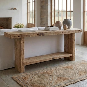 Peut inclure: Une console en bois rustique avec une étagère inférieure. La table est en bois vieilli et a une finition naturelle. Des vases décoratifs et des livres sont exposés sur la table. Un tapis tissé avec un motif géométrique est posé sur le sol.