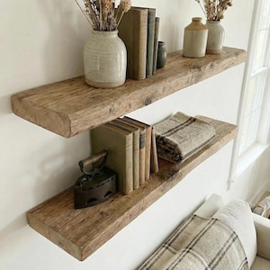 May include: Two rustic wooden floating shelves on a white wall. The top shelf has a ceramic vase with dried flowers and old books. The bottom shelf displays more books, a vintage iron, and a folded blanket.