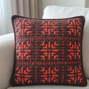 May include: A square crochet pillow with a geometric pattern in shades of brown and orange. The pillow features a detailed, textured design, with a dark brown border. The decorative pillow is placed on a light-colored sofa.