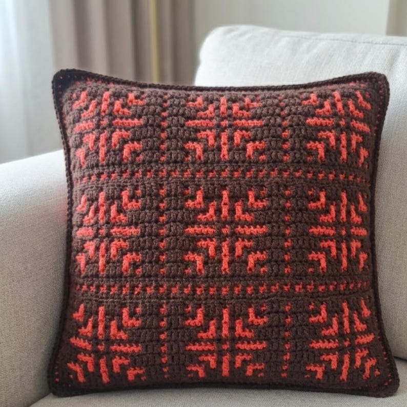 May include: A square crochet pillow with a geometric pattern in shades of brown and orange. The pillow features a detailed, textured design, with a dark brown border. The decorative pillow is placed on a light-colored sofa.