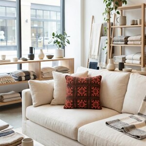 May include: A cream-colored sofa with several pillows, including a decorative square pillow with a brown and red geometric pattern. Shelves and a wooden ladder display folded blankets and decorative items. The room has a neutral color palette.