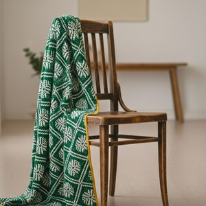 May include: A green and white patterned blanket draped over a wooden chair. The blanket has a repeating geometric design with a yellow border. The chair is made of dark wood and has a slatted back.