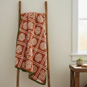 May include: A terracotta-coloured blanket with a white floral pattern and a green border is draped over a wooden ladder. The blanket's design features a repeating geometric pattern of squares with floral motifs.