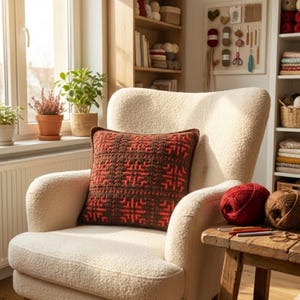 May include: A cozy cream-colored armchair with a brown and red patterned crochet pillow. A small wooden table holds red and brown yarn balls and crochet hooks. The scene is set in a well-lit room with plants and a bookshelf.